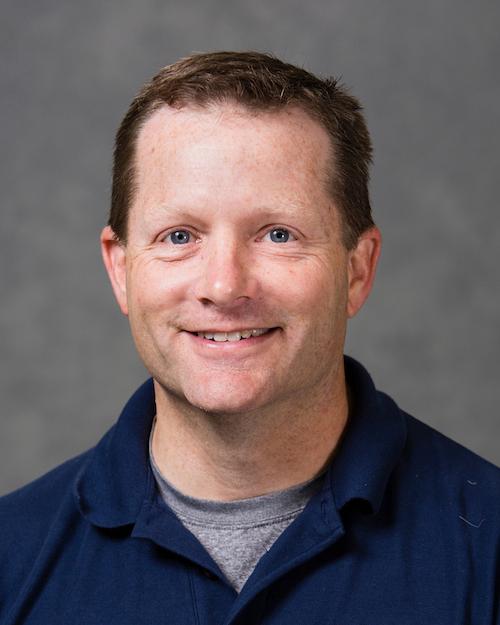 a smiling person with short brown hair wearing a navy blue shirt