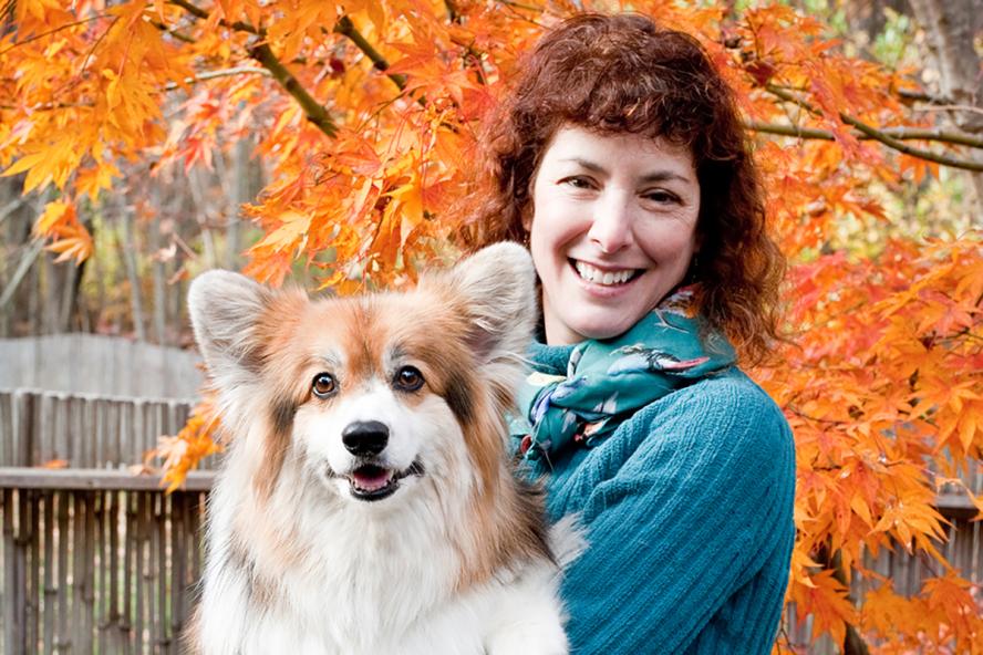 A smiling person with red curly hair holding a sheltland sheepdog. They are standing outdoors in the fall.