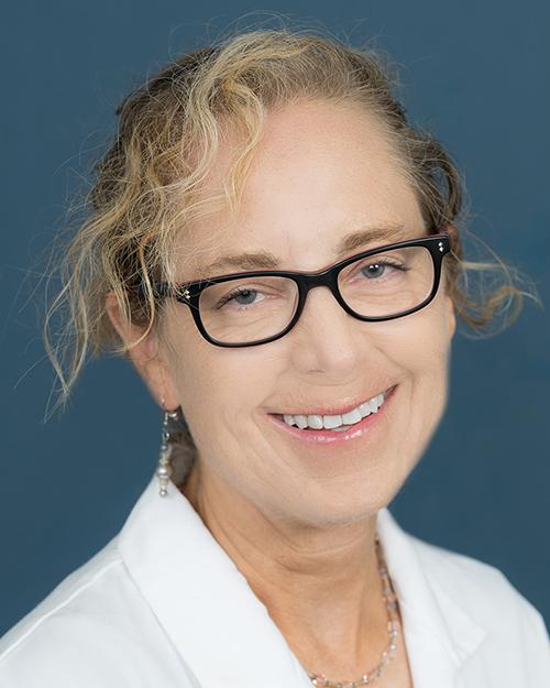 a smiling person with blong curly hair pulled back from face, wearing black eye glasses and a white medical coat