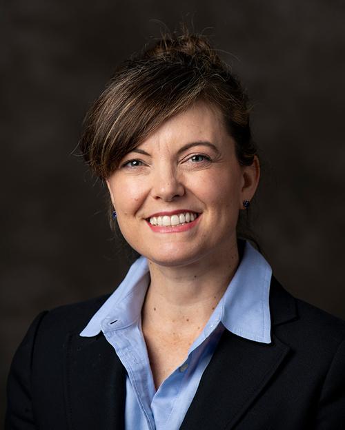 a smiling person with dark brown hair pulled up in a bun with bangs. They are wearing a blue button down shirt and navy suit jacket