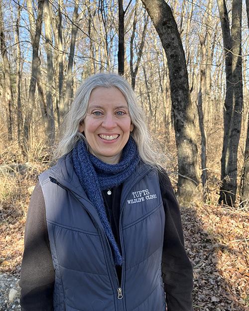 a smiling person with long grey hair wearing a black long sleeve shirt and navy blue vest standing in the woods.