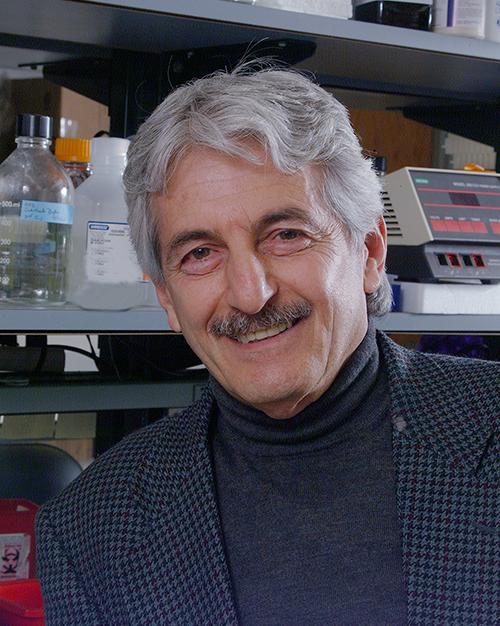 a smiling person with grey hair and a mustache wearing a turtle neck sweater and suit jacket. He is in a lab.