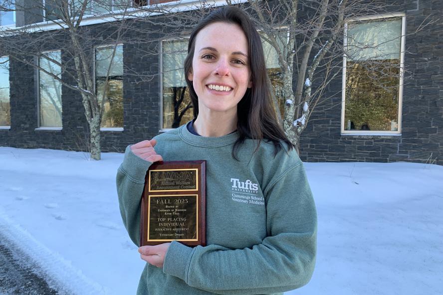 a smiling individual with long brown hair wearing a green sweatshirt holds an award