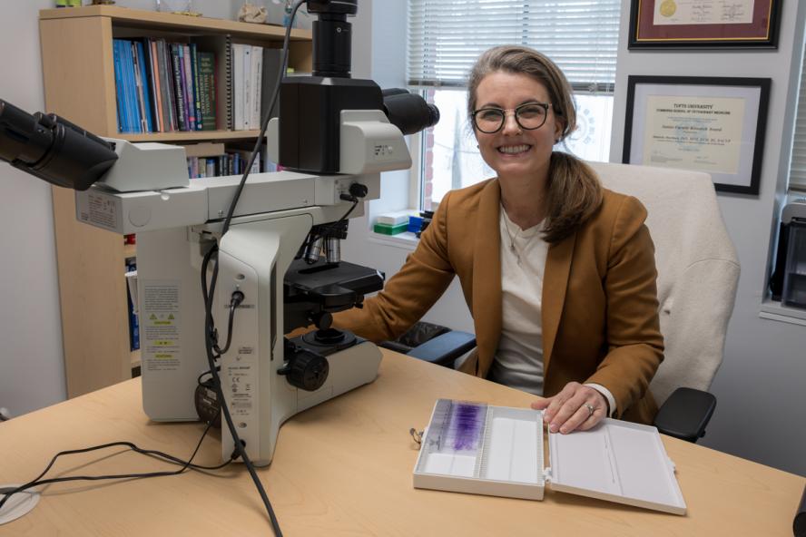 A smiling person with long hair and glasses sitting at a desk with a microscope.