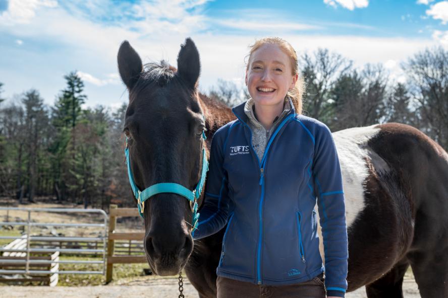 A smiling person smiling wearing ith a blue coat standing next to a black horse outside.