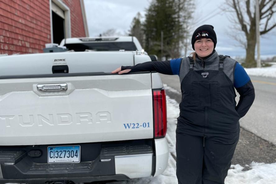 Person smiling with a black hat on and dark overalls standing next to a white truck outside in the cold.