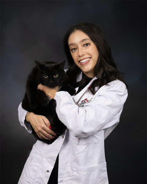 a smiling indiviudal with shoulder length black hair wearing a white medical coat and holding a black cat.