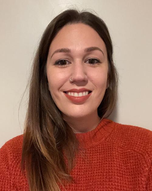 A smiling person with long light brown hair hanging over one shoulder. They are waring a dark orange sweater