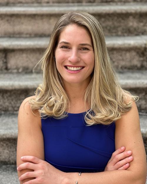 A smiling individual with shoulder length blond hair wearing a royal blue top and has their arms crossed.