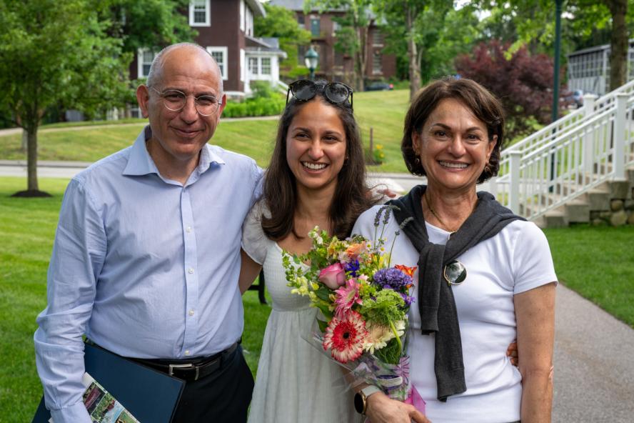 Three smiling people, one holding flowers, standing together.