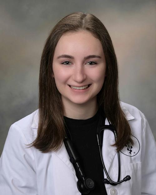 a smiling individual with shoulder length brown hair wearing a black shirt, white medical coat and a stethoscope around their neck