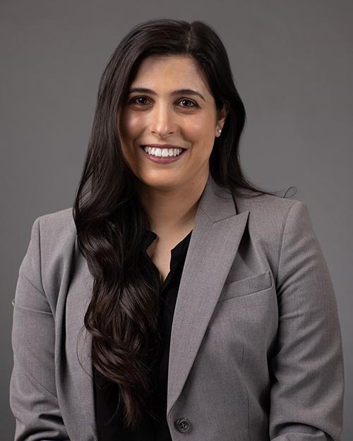 A smiling individual with long black hair wearing a black blouse and gray suit jacket.