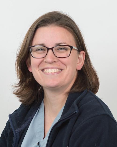 A smiling person with brown shoulder-length hair, wearing glasses and a navy blue polar fleece jacket.