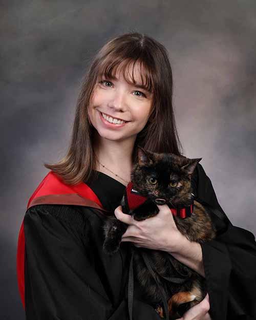 A smiling person with long brown hair wearing a black and red graduation robe and they are holding a cat.