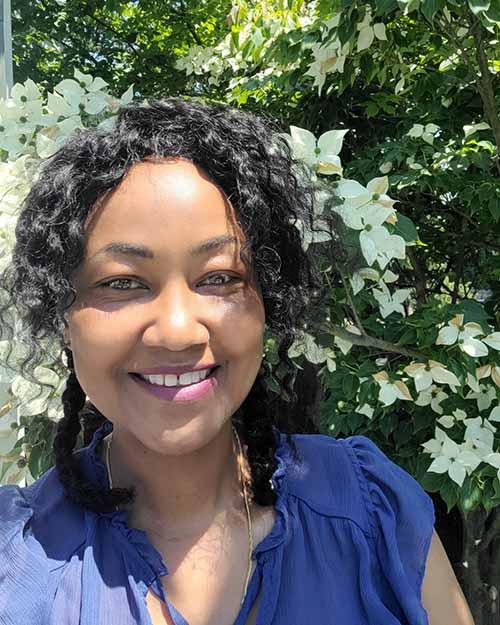 a smiling person with black curly hair wearing a blue sleeveless blouse.