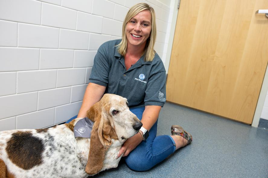 Smiling person with shoulder length blond hair sitting on the ground of an exam room floor, holding Bubba the Basset Hound in their lap.