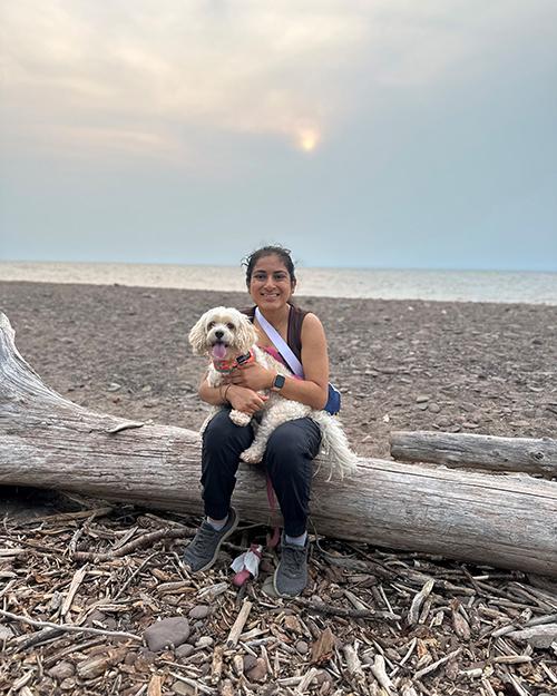 a smiling individual sitting on a dead tree stuck on a beach. They are holding a small white dog.