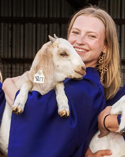 A smiling individual with strawberry blond hair wearing a blue graduation gown and holding a baby goat.