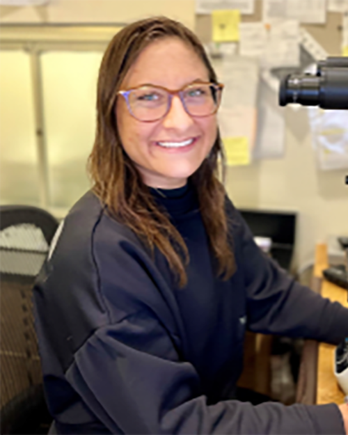 A smiling person with long brown hair wearing glasses and a long sleeve navy blue shirt.