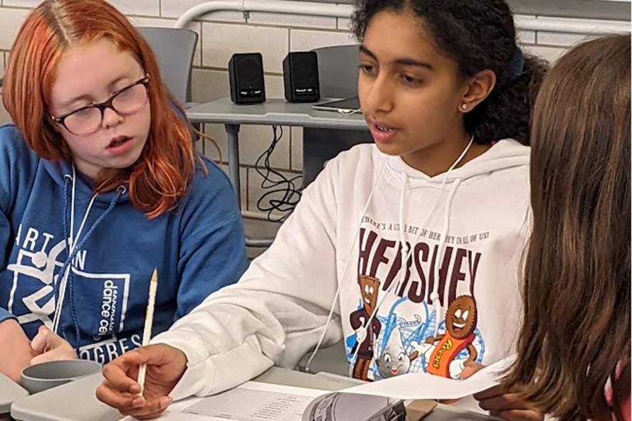 Two students in a classroom setting, one with glasses wearing a  blue sweatshirt and the other student holding a pencil and a clipboard with black hair tied back and a white sweatshirt with brown lettering on it.