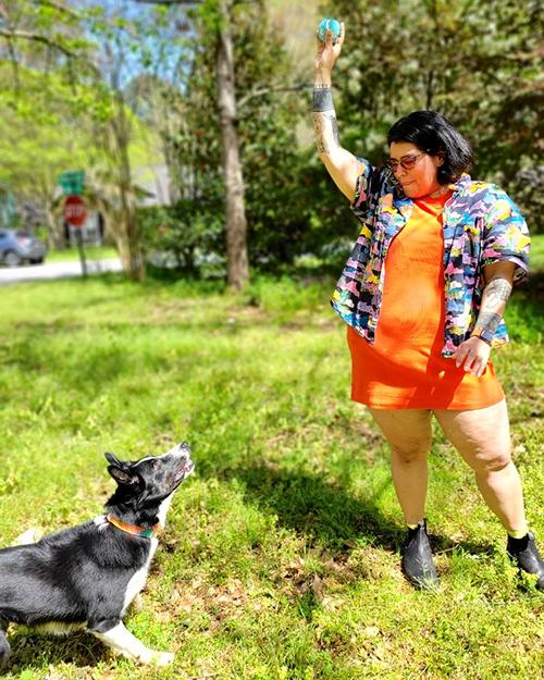 a person with black hair, wearing glasses, a short orange dress and multicolored short sleeve blouse and black booties. They are playing ball in a park with a black and white dog.