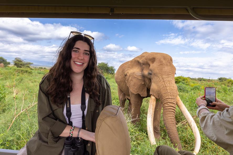 Person smiling with binoculars around neck, and an elephant with large tusks in background.