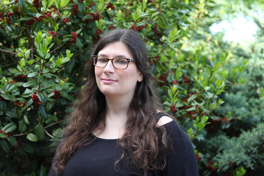 an individual with long dark hair wearing glasses and a black shirt standing outdoors in front of a green bush with red berries.