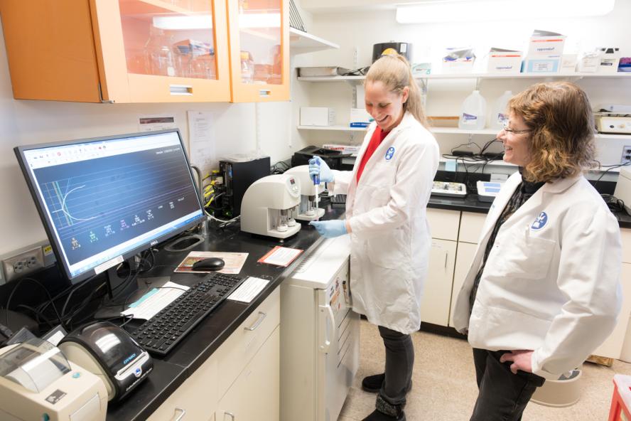 Two smiling individuals interacting in a lab and both wearing white lab coats.