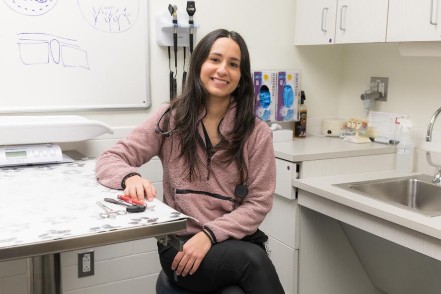 A person with long dark hair sits smiling in an exam room with black pants and a mauve top on.