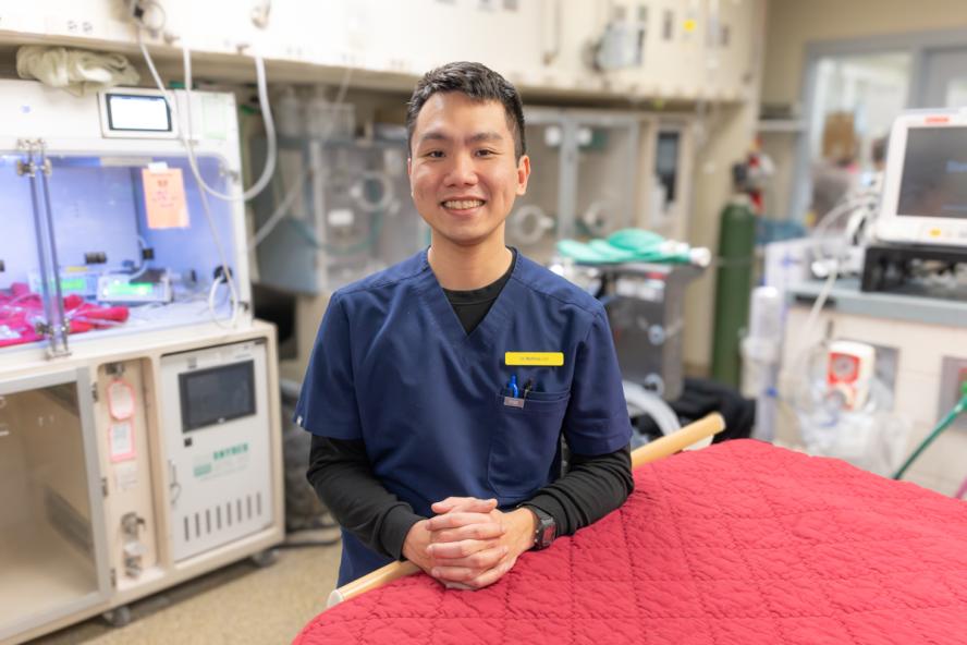 Person smiling with short black hair wearing a blue scrub shirt in a medical room.