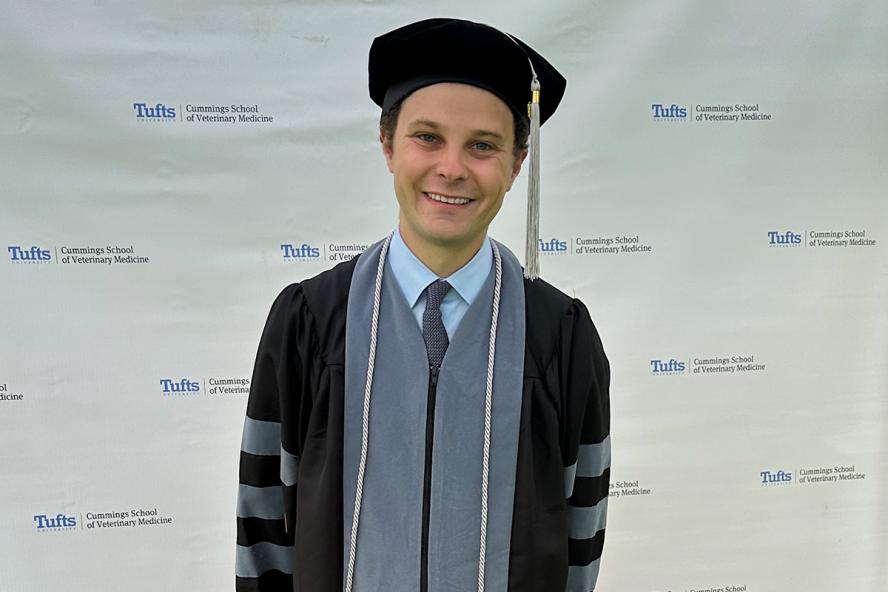 Person smiling wearing a cap and gown standing in front of a backdrop titled  Cummings School of Veterinary Medicine at Tufts University.