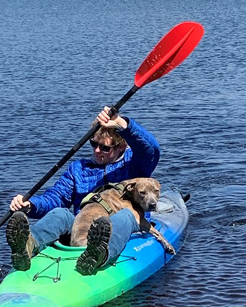 a person wearing jeans, blue jacket and sunglasses is kayaking with their dog.