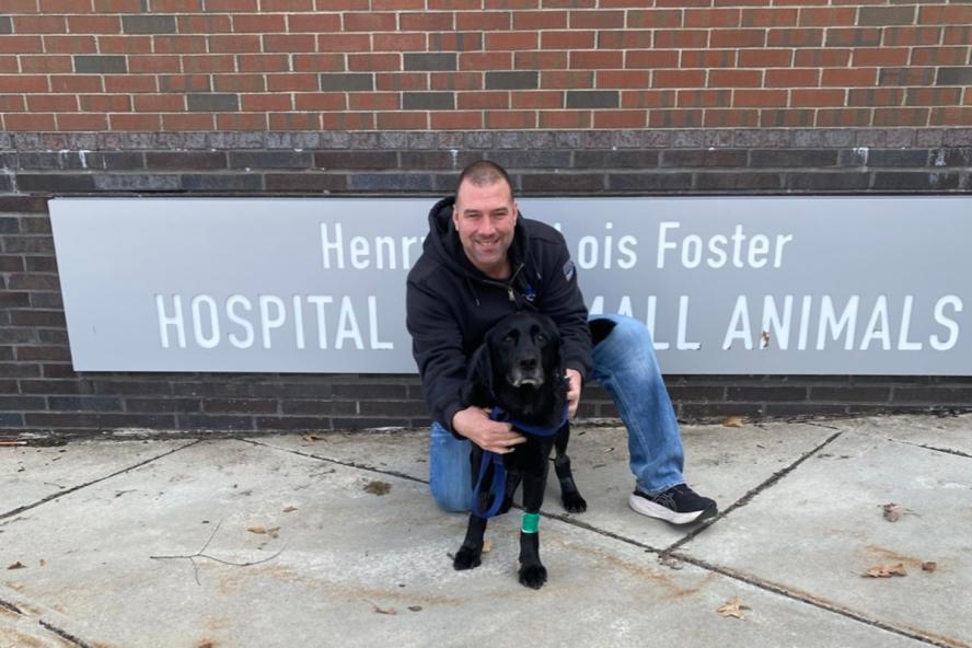 Black Labrador stands outside of Foster Hospital for Small Animals at Cummings School of Veterinary Medicine with the owner holding him.