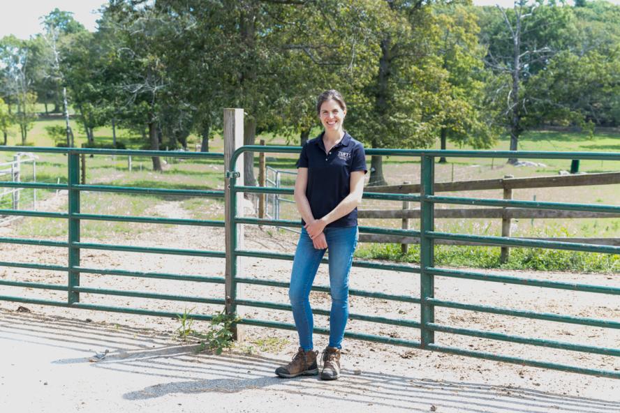 Person standing outside in front of a cattle gate and farm field. 