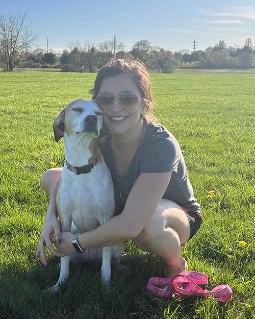 Andrea Rocha bent down hugging her white and brown dog in a field of green grass.