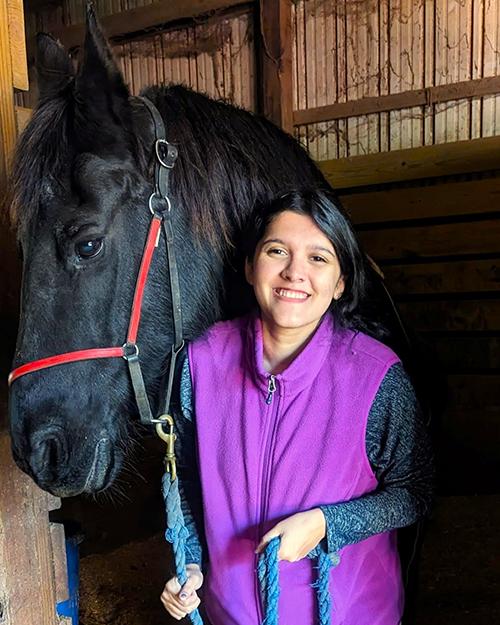 Elizabeth Singh smiling, wearing a purple vest and standing next to a black horse in a barn.
