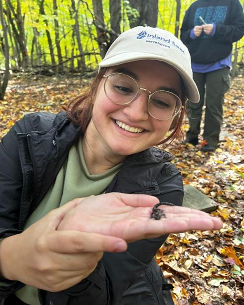 Kendall Kindzierski is smiling, wearing glasses and a cap and is standing in the woods holding a small lizard in her hand.