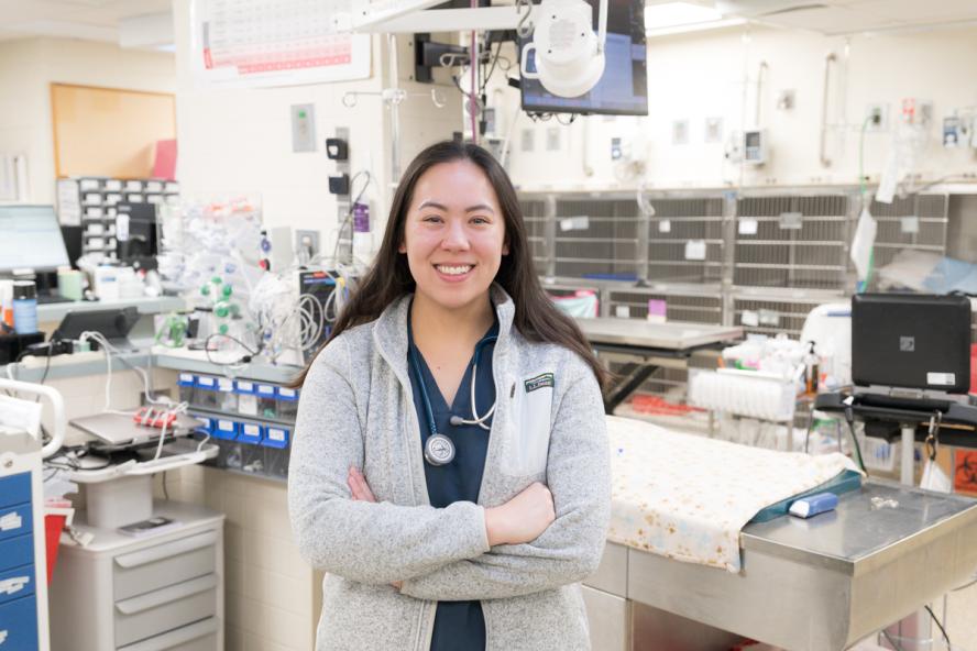 Smiling veterinary professional wearing scrubs and a stethoscope stands with arms crossed in a brightly lit veterinary treatment room filled with medical equipment, exam tables, and monitors.