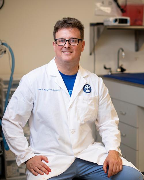A veterinarian, wearing a white medical coat, glasses, and a smiling face, sits in a chair in an exam room.
