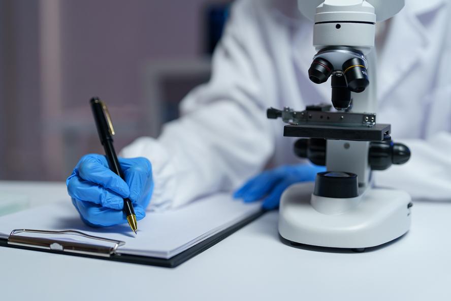 Close-up of scientist's hands in blue gloves writing notes on clipboard while conducting research using a microscope in a modern laboratory setting, emphasizing scientific analysis and discovery. Adobe Stock/Phimwilai