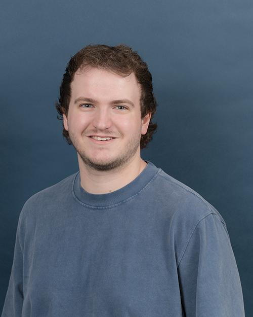 Dylan Thomas has short brown wavy hair, is smiling and wearing a blue-grey shirt.