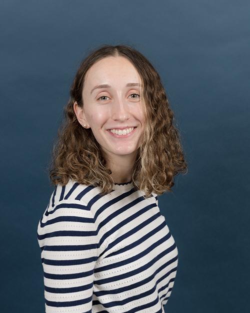 Emma Riguad has wavy shoulder length light brown hair, is smiling and wearing a black and white striped long sleeve shirt.