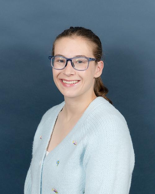 Leah Fine smiling, wearing blue glasses and a light blue sweater. She has her hair pulled back in a pony tail.