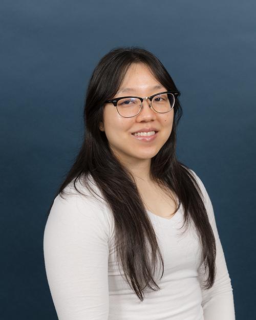 Lira Bouthsarath has long black hair, is smiling, wearing glasses and has a white long sleeve shirt on.