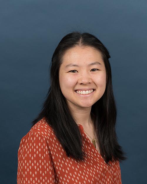 Alyssa Dalrymple has medium length black hair parted in the middle. She is smiling and wearing an orange colored geometric design blouses.
