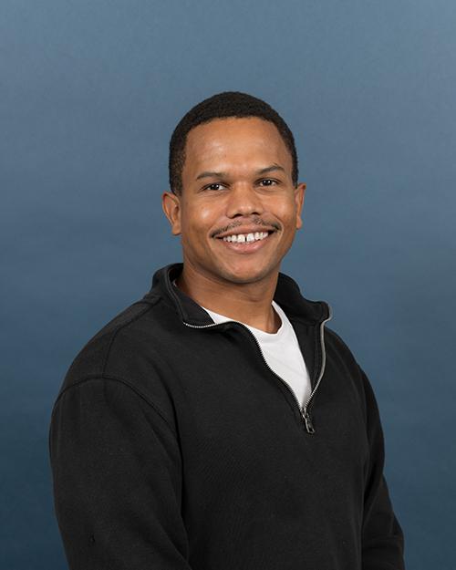 Damian Clarke has very short black hair, is smiling and wearing a white shirt with a long sleeve quarter zip black pullover.