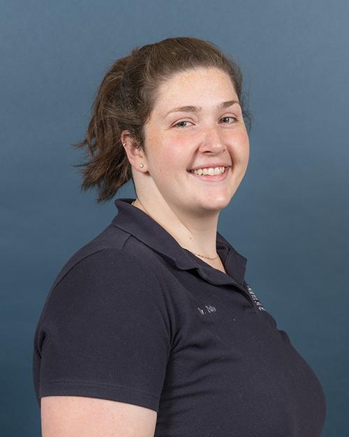 Kim Zullo has medium length brown hair pulled back in a pony tail. She is smiling and wearig a navy blue short sleeve polo shirt.