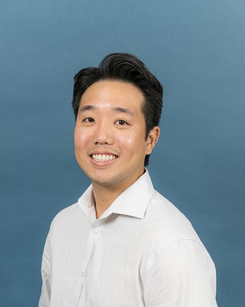 Zachary Low has short wavy black hair, is smiling and wearing a white button down long sleeve shirt.
