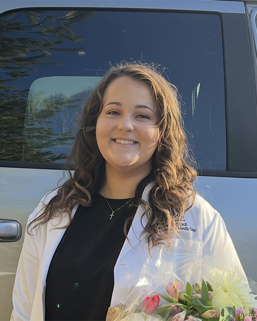 Nicole Tiberio smiling, has long brown wavy hair, is wearing a white medical coat, and is standing outside in front of a truck.