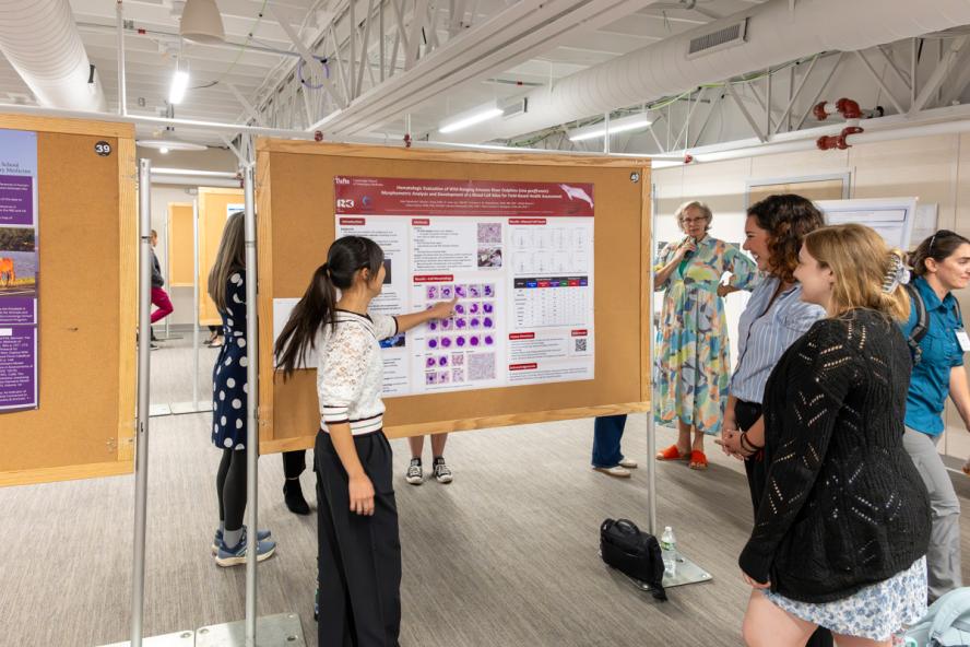 A student points to a poster with graphics and text while attendees look on and observe  other posters.